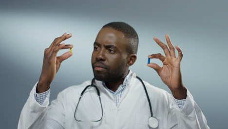 African-American-young-male-physician-holding-two-pills-in-hands,-one-yellow-and-one-blue,-while-comparing-them-and-studying-carefully.-Close-up.